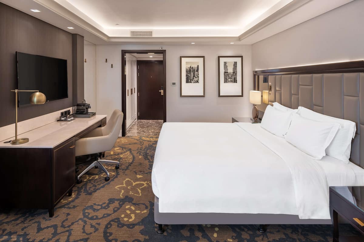 A hotel room with a large bed, a desk with a flat-screen TV, a chair, and two framed pictures on the wall.