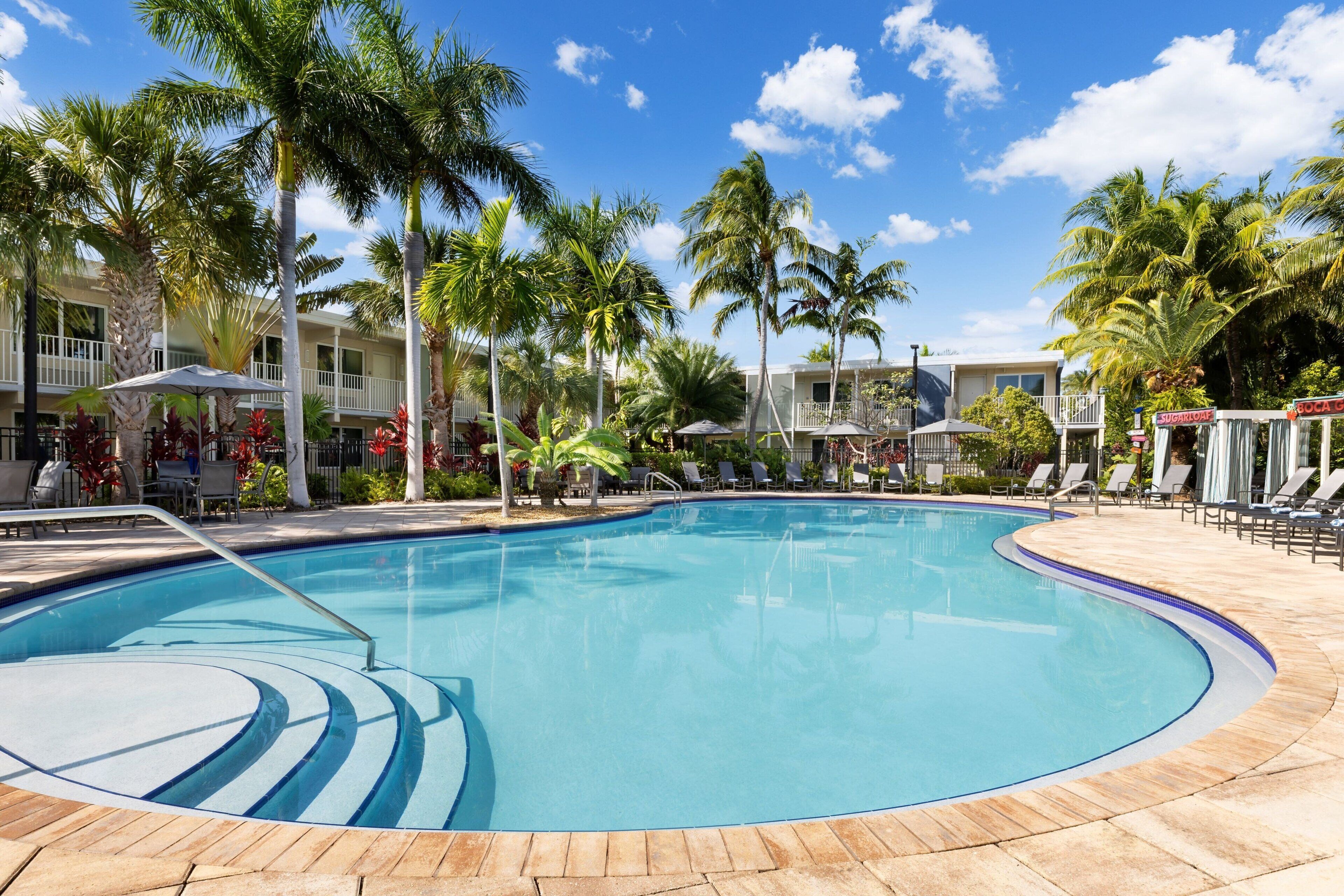 A pool area with lounge chairs and umbrellas, surrounded by palm trees and a building with balconies.