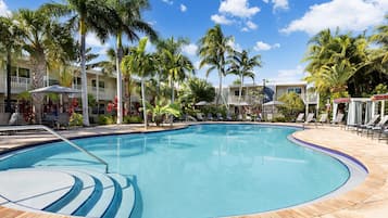 A pool area with lounge chairs and umbrellas, surrounded by palm trees and a building with balconies.