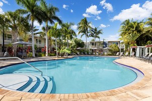 A pool area with lounge chairs and umbrellas, surrounded by palm trees and a building with balconies.