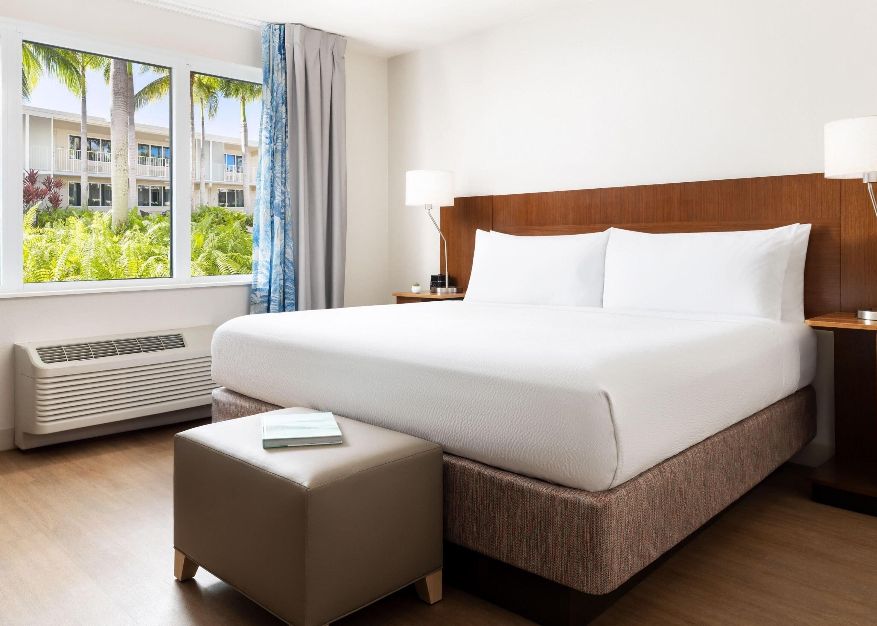 A hotel room with a large bed, a bedside table with a lamp, a window with curtains, and a view of greenery outside.
