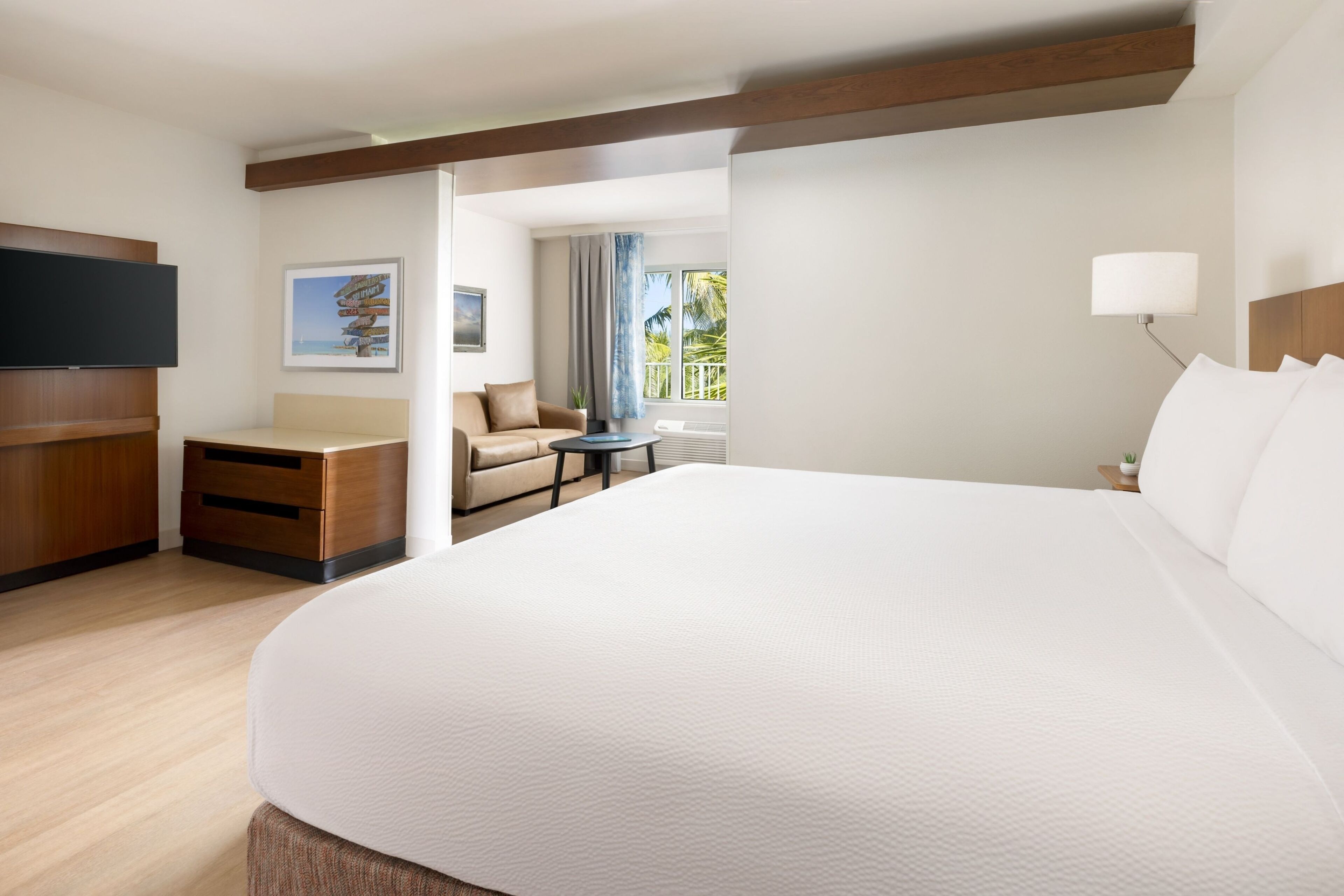 Une chambre d’hôtel avec un grand lit, un téléviseur à écran plat, un canapé, une petite table et une fenêtre donnant sur des palmiers.