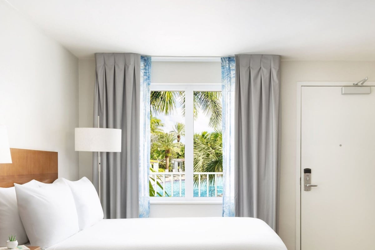 A hotel room with a bed, a window with curtains, a view of a pool and palm trees, and a door with a handle.