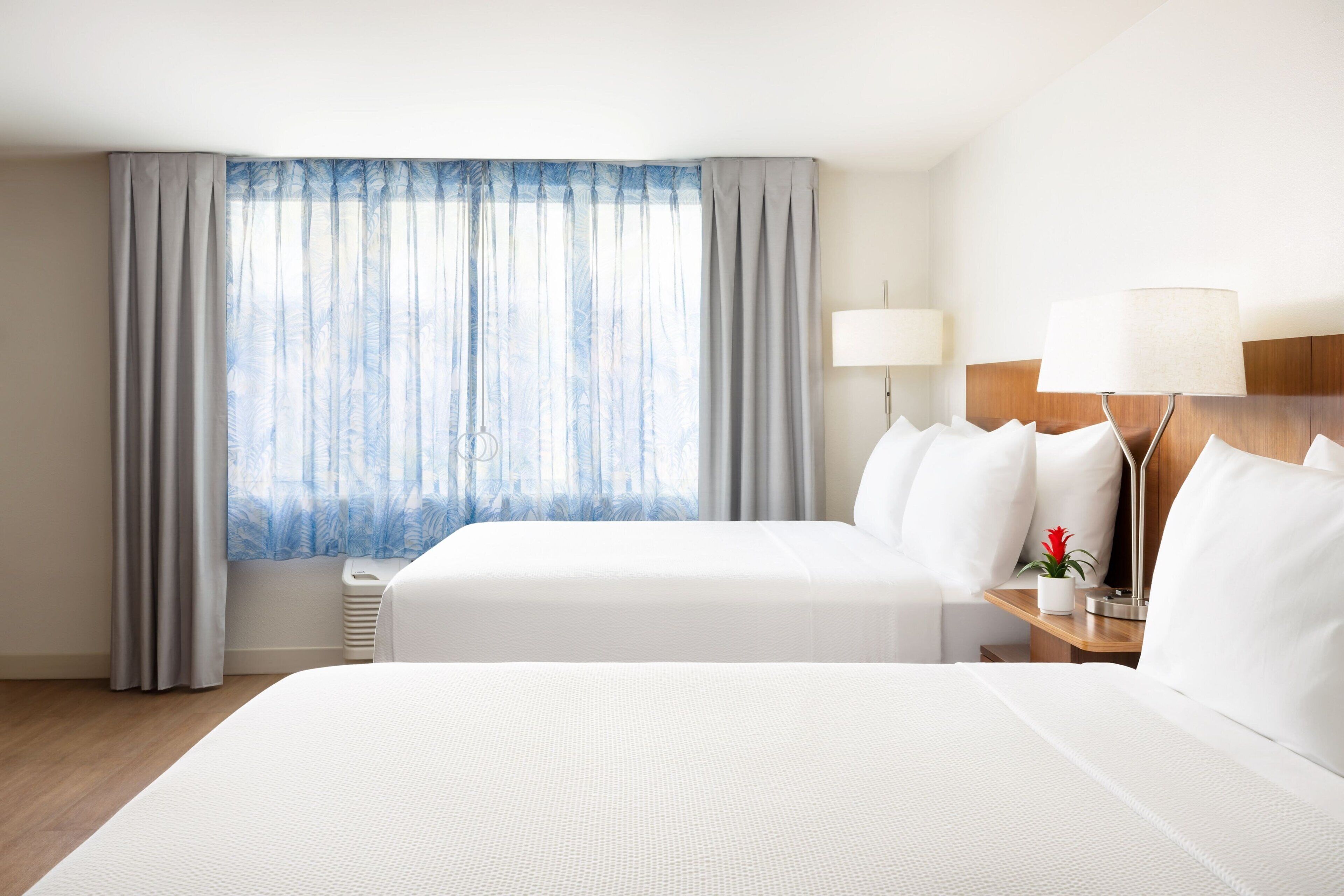 Une chambre d’hôtel avec deux lits, une table de chevet avec une lampe, une fenêtre avec des rideaux et une petite table agrémentée d’un vase de fleurs.