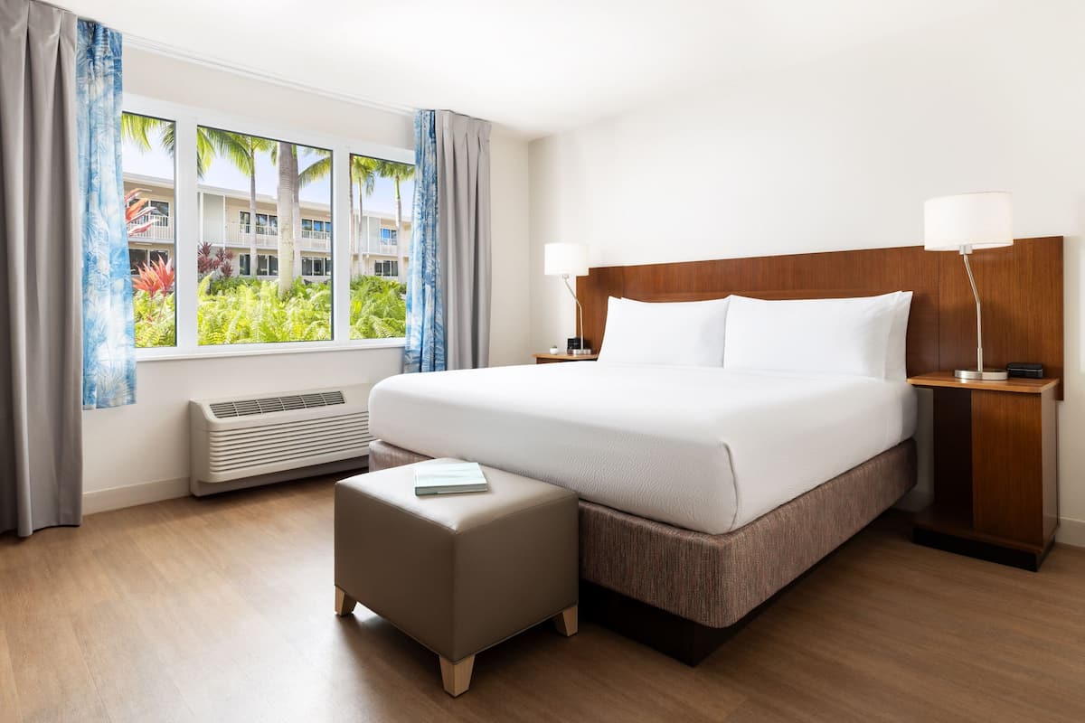 A hotel room with a large bed, a bedside table with a lamp, a window with curtains, and a view of palm trees.