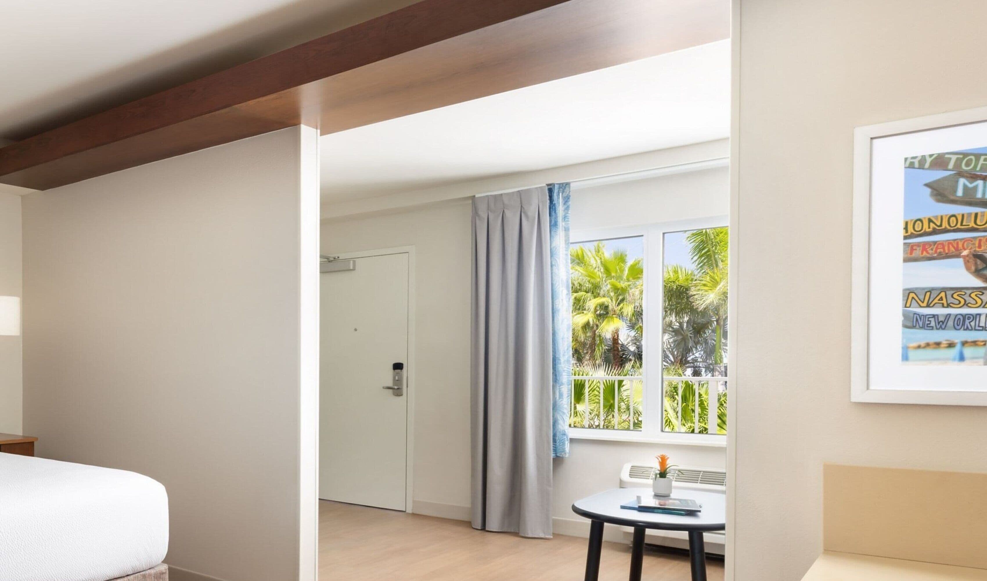 A hotel room with a bed, a small table with a plant, a window with a view of palm trees, and a framed sign on the wall.