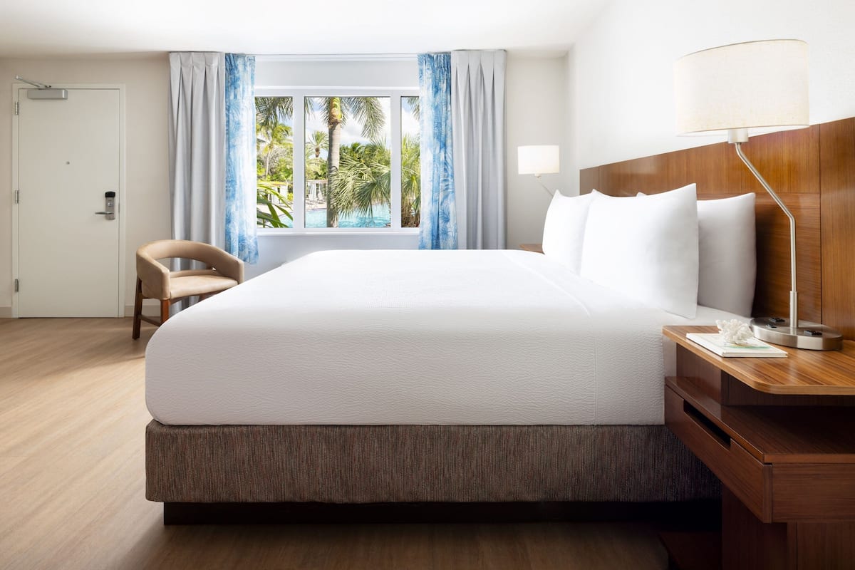 A hotel room with a large bed, a wooden nightstand, a desk, a chair, and a window with curtains.