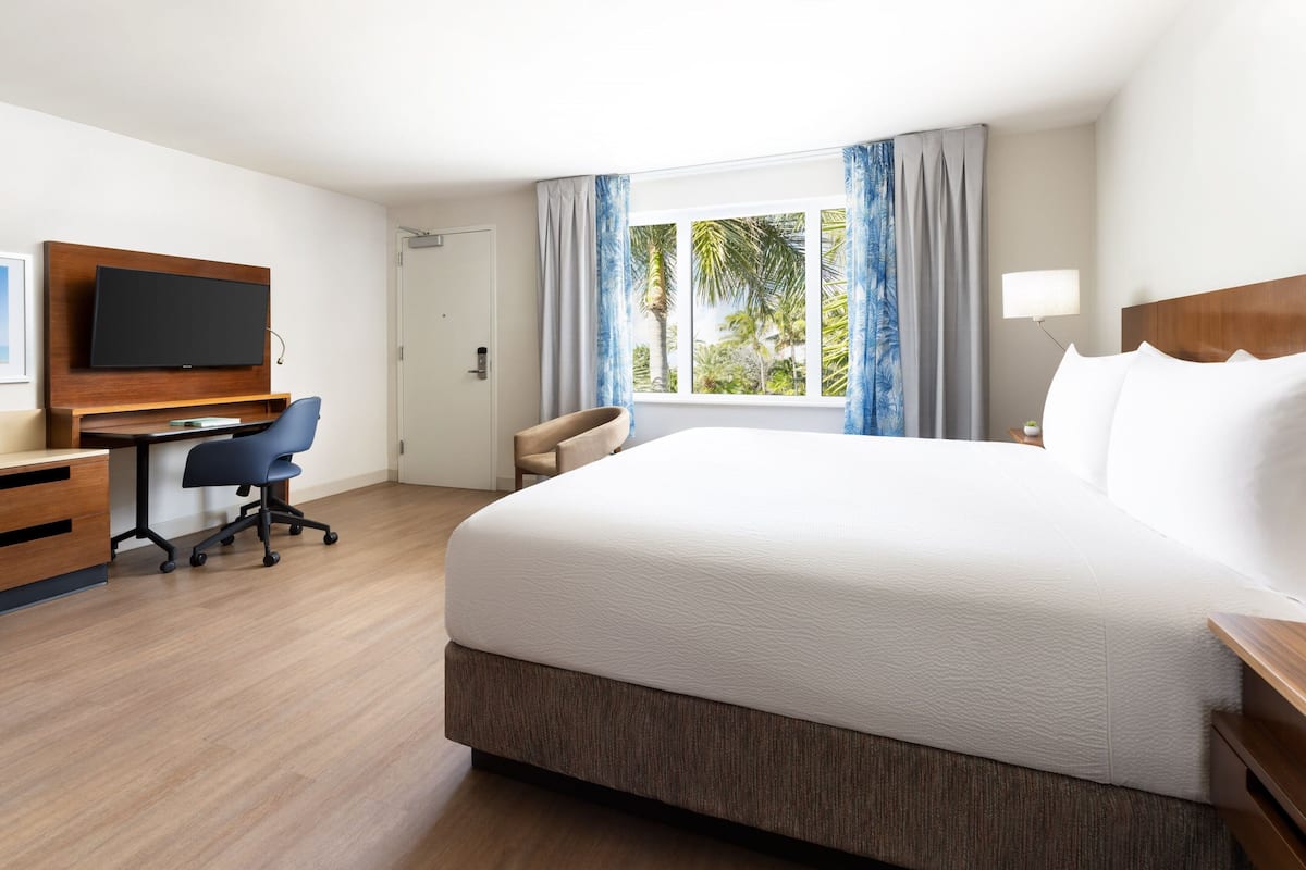 A hotel room with a large bed, a desk with a chair, a flat-screen TV, and a window with curtains.