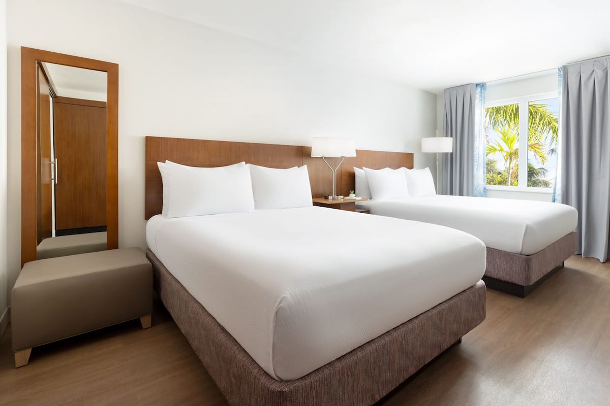 A hotel room with two beds, a wooden headboard, a nightstand with a lamp, a bench, and a window with curtains.