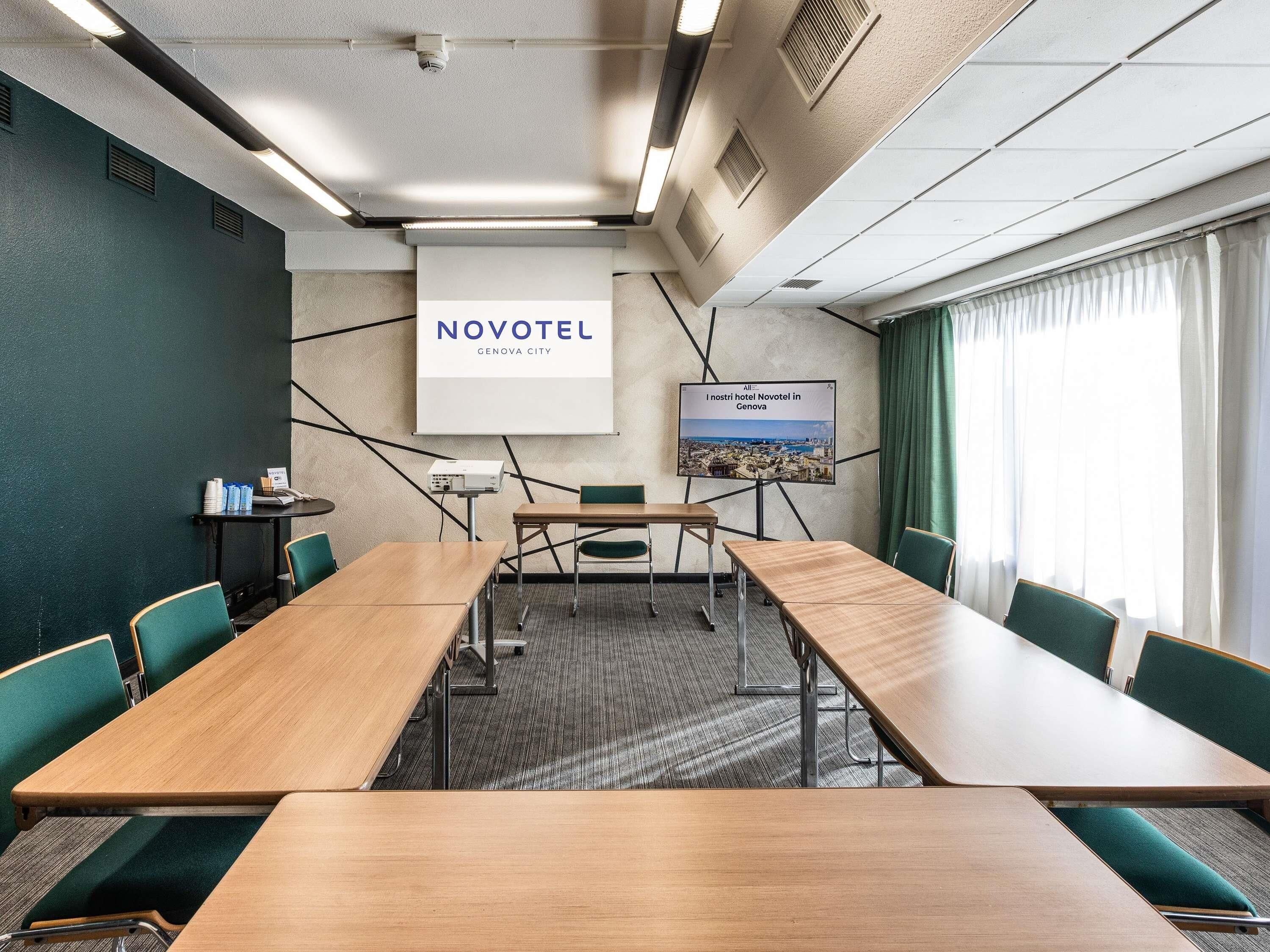Une salle de conférence équipée d’une longue table, de chaises, d’un projecteur et d’un écran affichant le texte « NOVOTEL Genova City ».