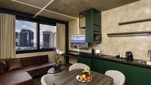 A modern hotel room with a dining area, kitchenette, and a view of the cityscape.