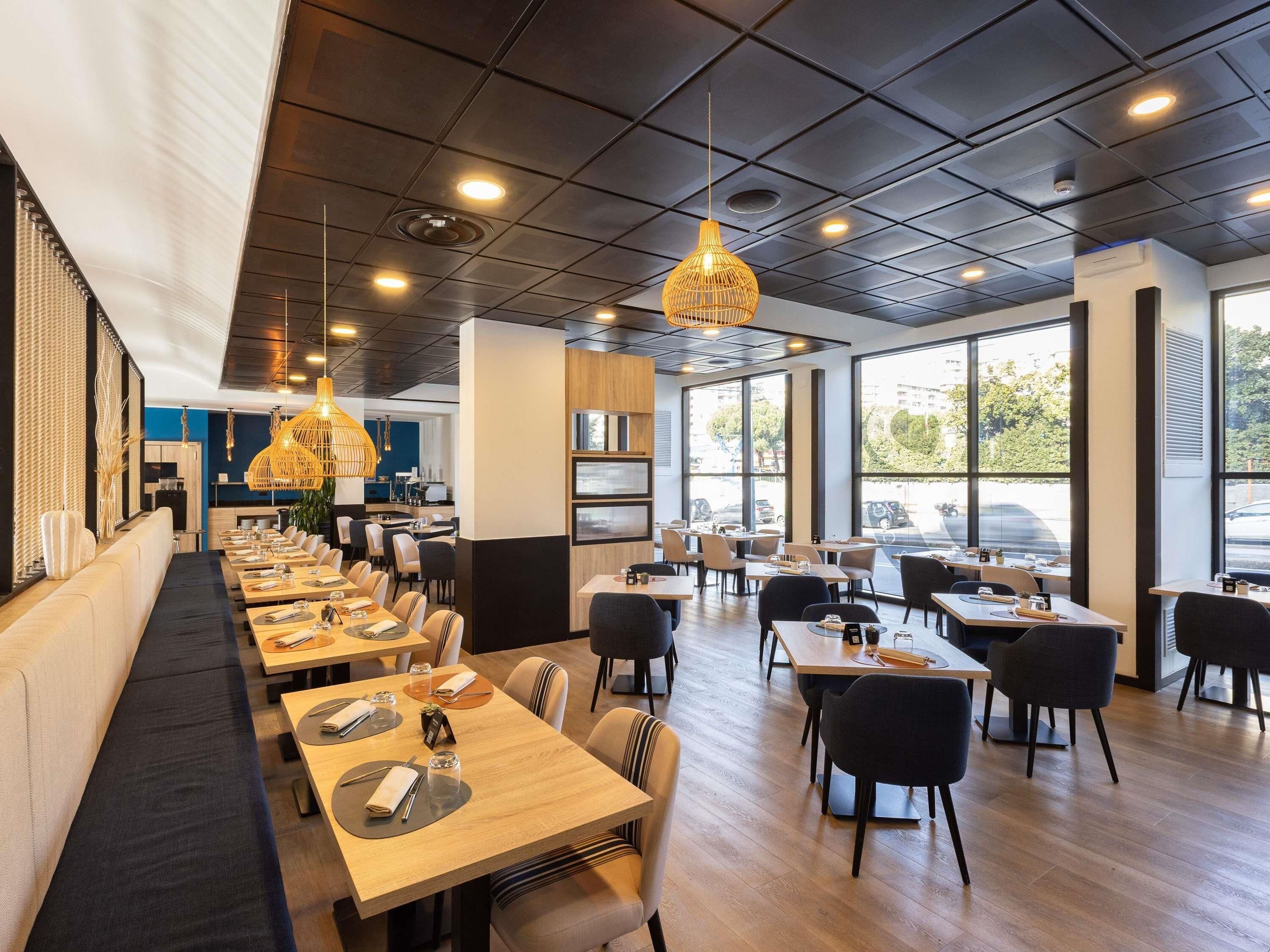 A modern restaurant with long communal tables, individual booths, and large windows offering a view of the street.