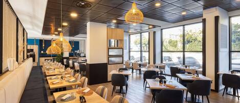 A modern restaurant with long communal tables, individual booths, and large windows offering a view of the street.