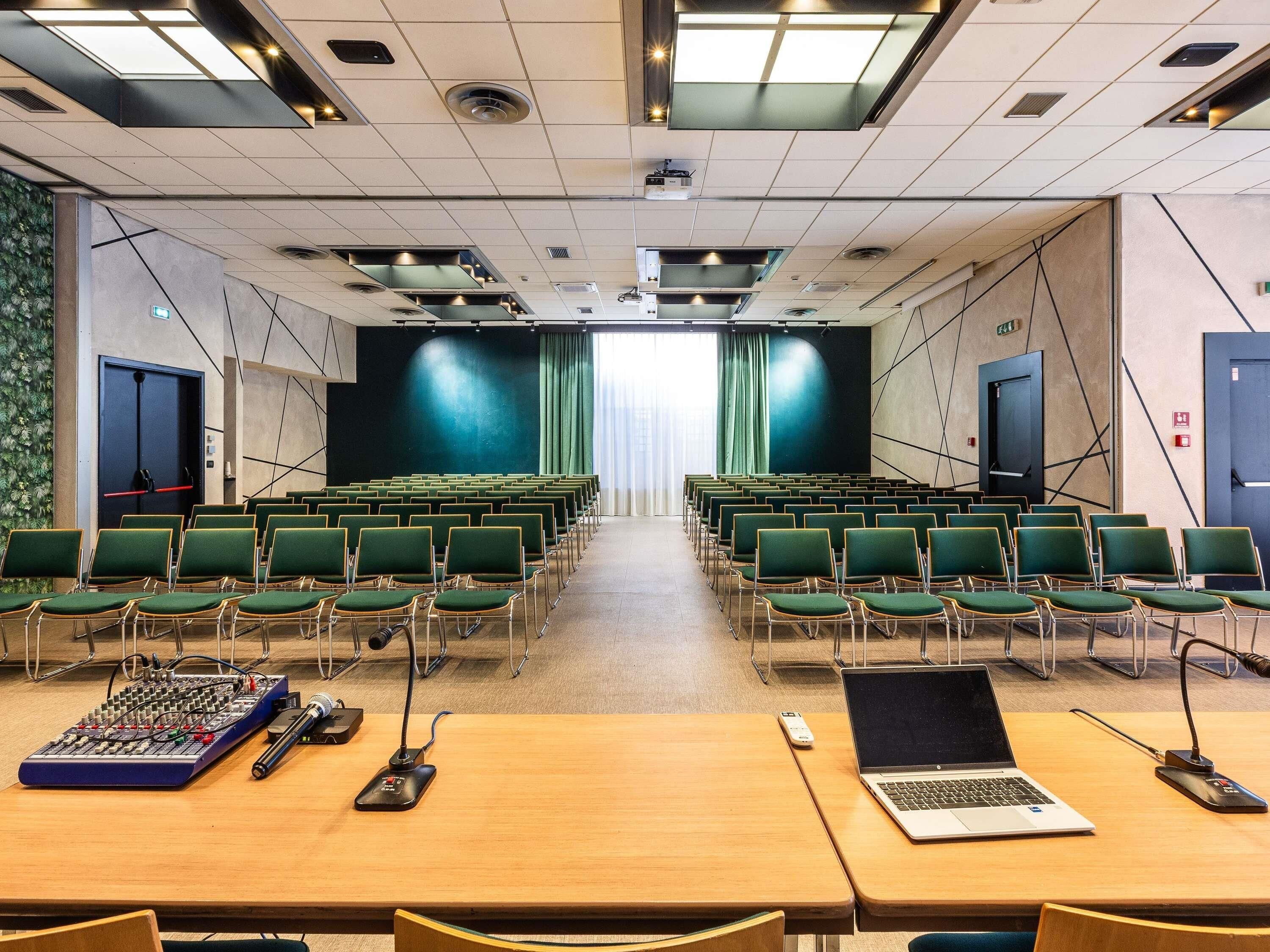 Une salle de conférence avec des rangées de chaises vertes, une scène dotée d’un fond noir, un projecteur et un ordinateur portable posé sur une table.
