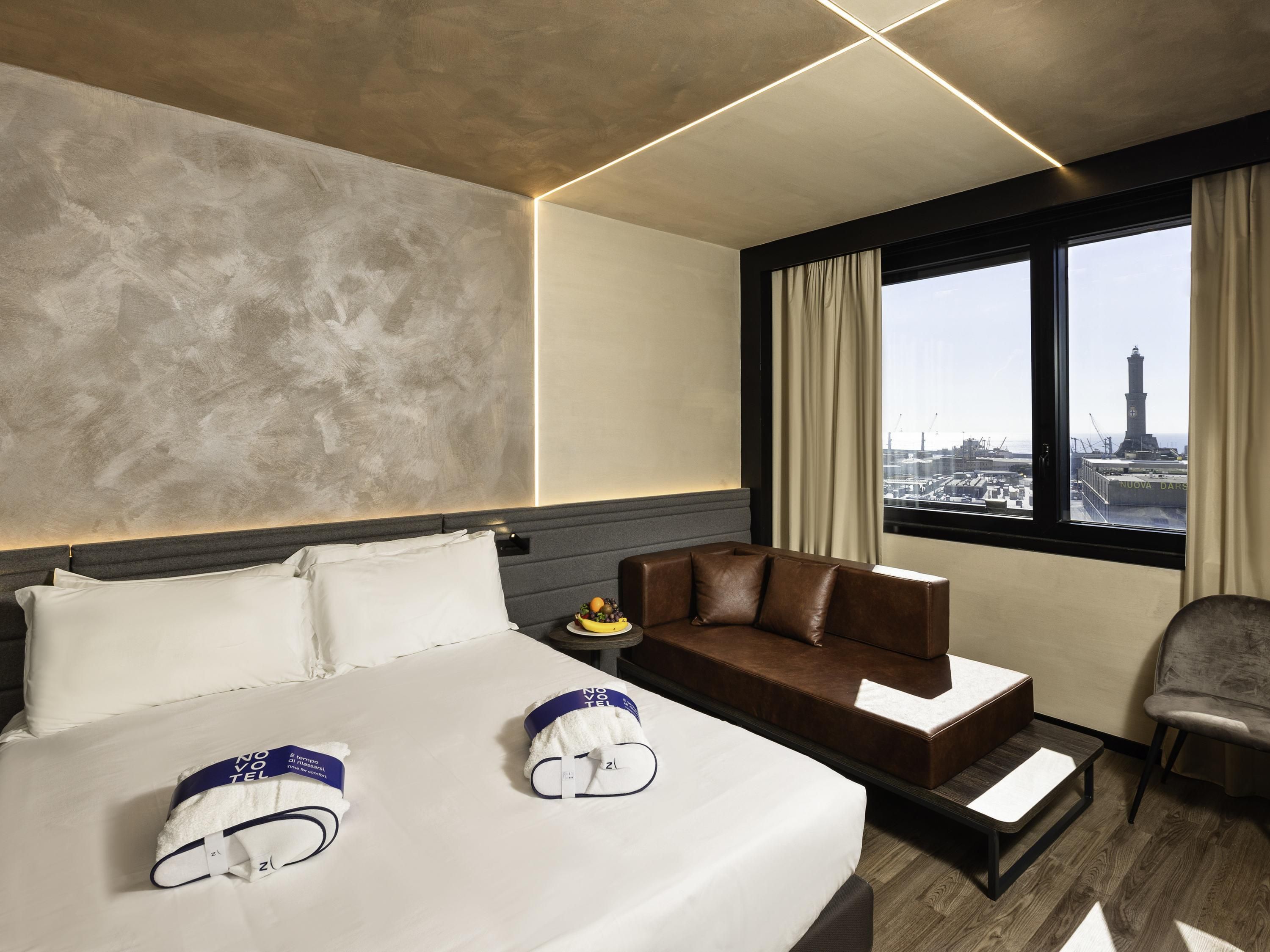 Hotelkamer met een bed, een bank, een stoel, een raam met uitzicht op een stadsbeeld, en een hoofdbord met een lichtbalk.
