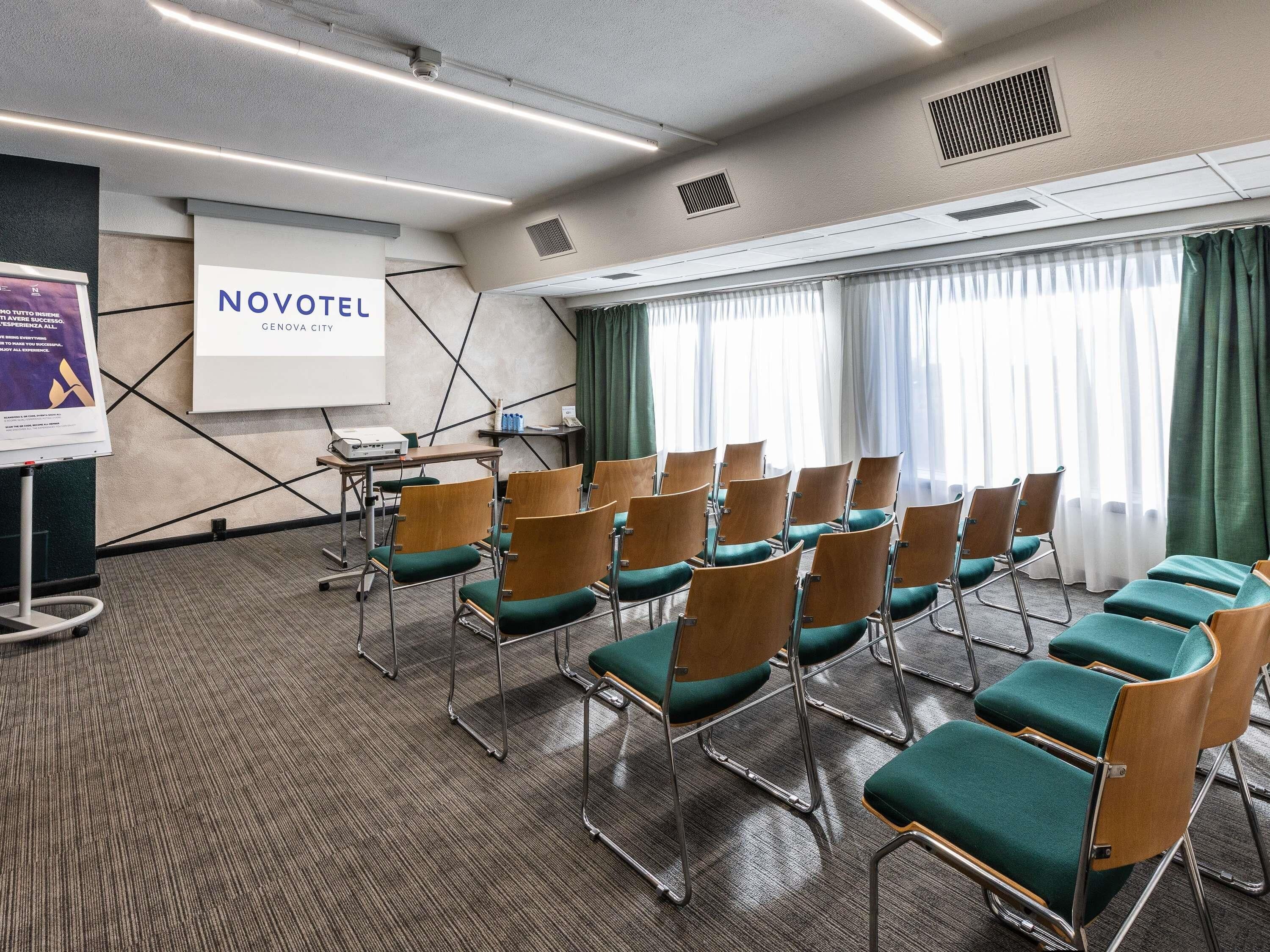 Een vergaderruimte met rijen stoelen, een projectiescherm en een bordje met Novotel Genova City.