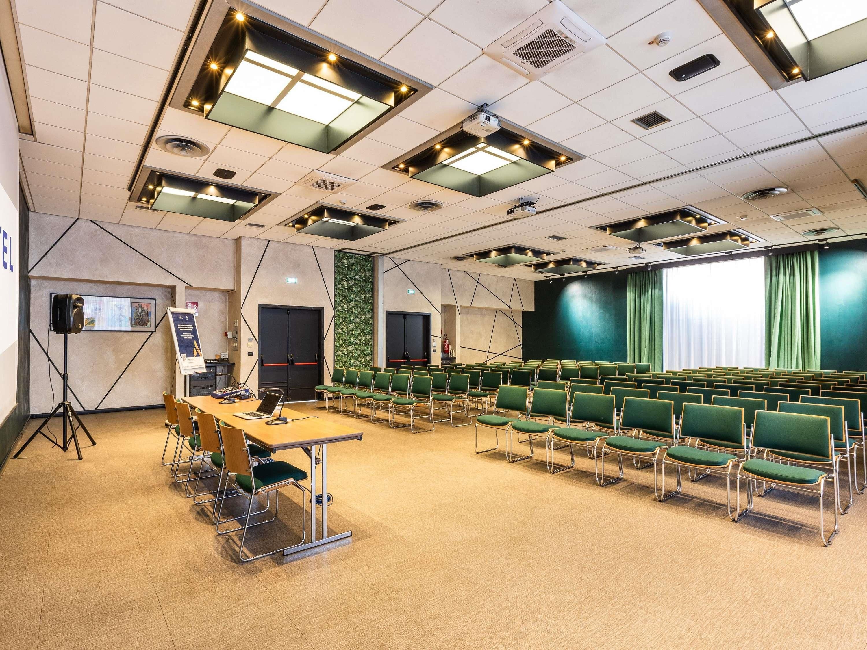 Une salle de conférence équipée d’une scène, de rangées de chaises vertes, d’un projecteur et d’un grand écran.