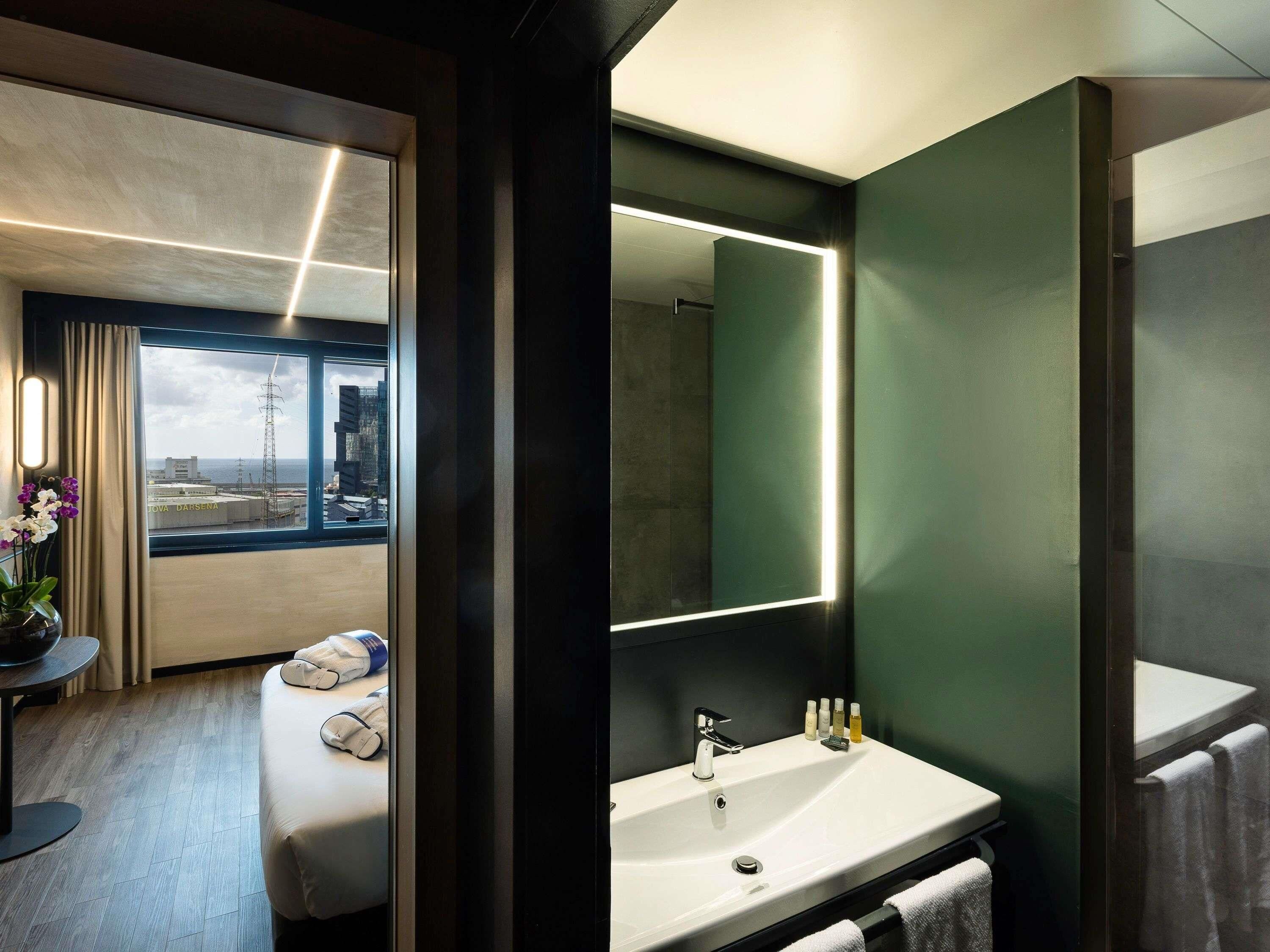 A modern hotel room with a large window, a round bed, a bathroom with a sink and mirror, and a view of the cityscape.