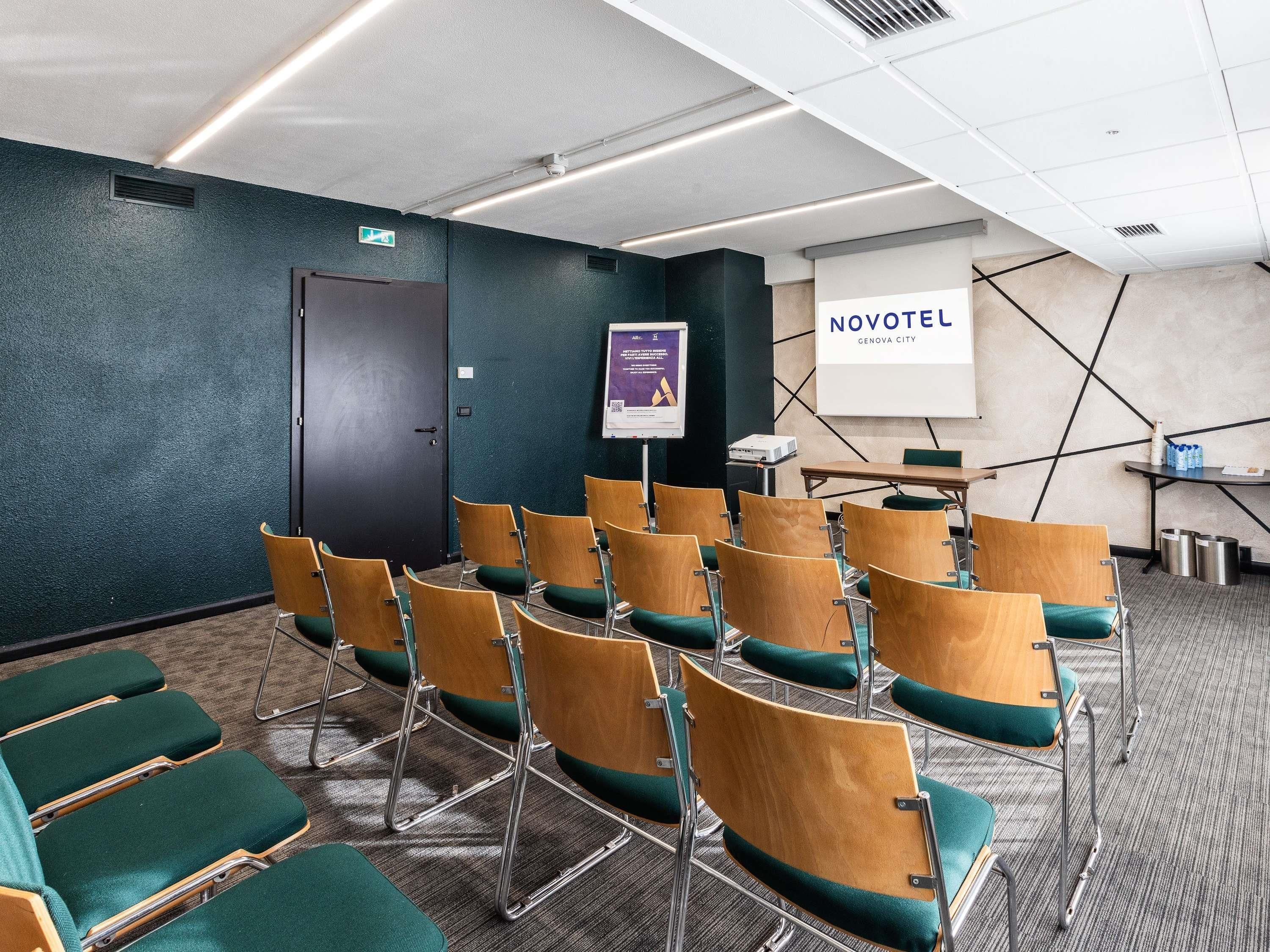 Una sala de conferències amb files de cadires de fusta amb coixinets verds, un projector, una pantalla i un rètol que diu 'NOVOTEL GENOVA CITY'.