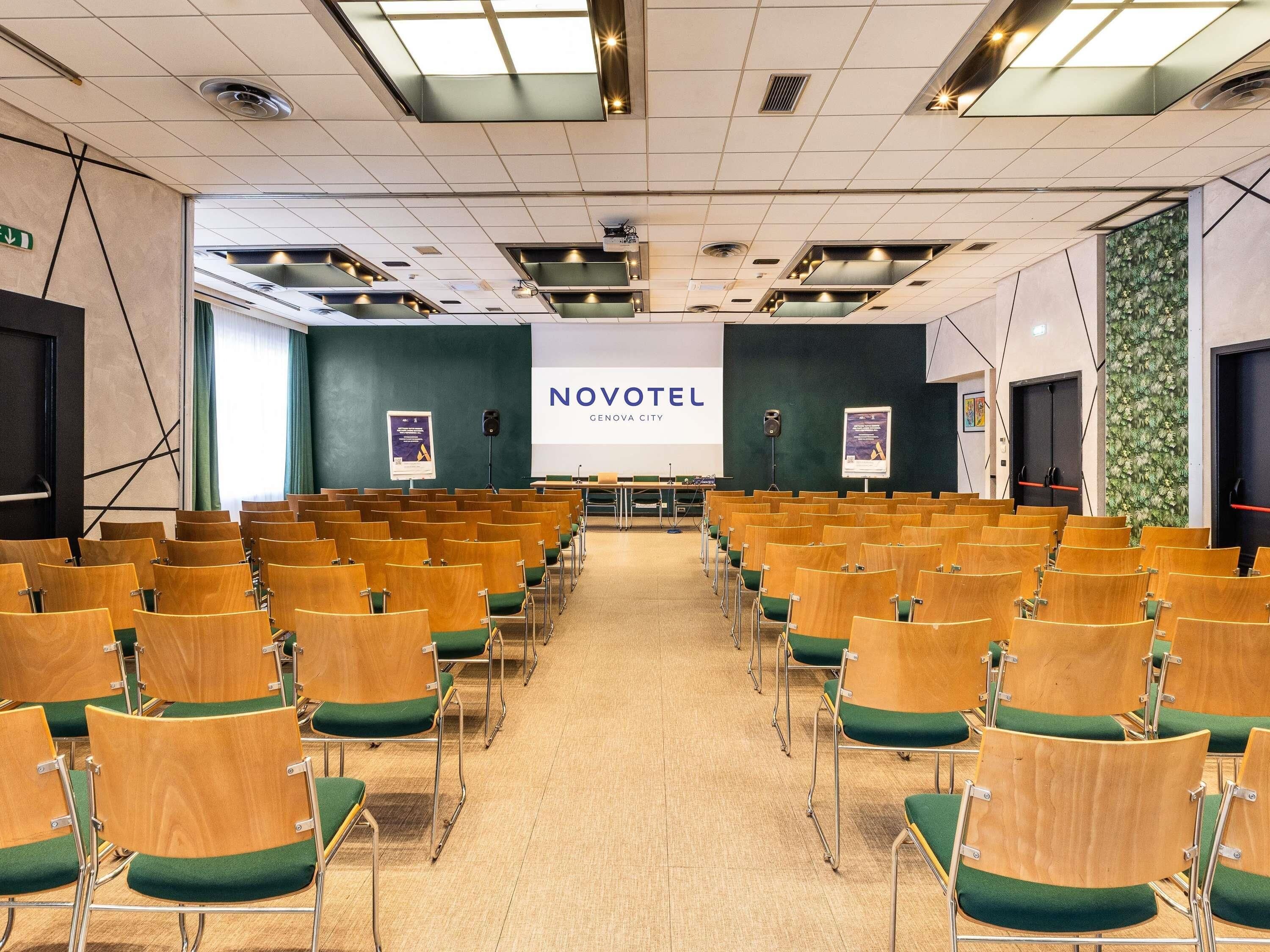 Een vergaderzaal met rijen houten stoelen en groene kussens, een groot scherm waarop 'NOVOTEL GENOVA CITY' staat, en een groene wand met een bloemistisch patroon.