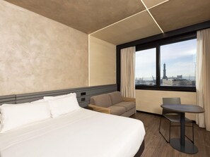 A hotel room with a bed, a sofa, a small table, and a window with a city view. - Novotel Genova City (Genoa)