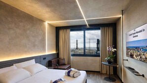 A hotel room with a large bed, a small seating area, a window with a city view, and a poster on the wall.