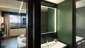 A modern hotel room with a large window, a round bed, a bathroom with a sink and mirror, and a view of the cityscape.