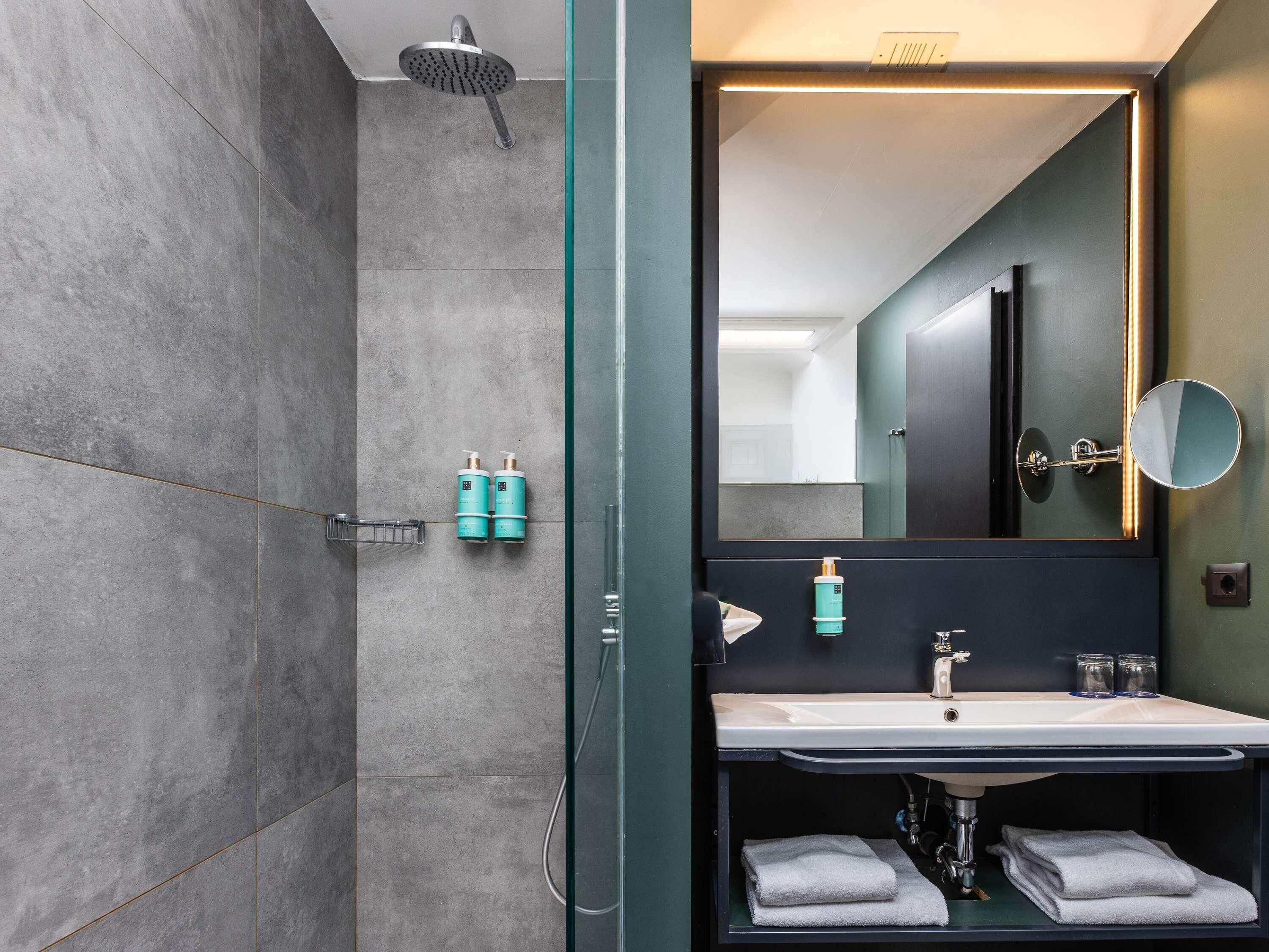 Ein modernes Badezimmer mit einer Glaskabine zur Dusche, einem großen Spiegel, einem Waschbecken und zwei Shampoo-Flaschen.