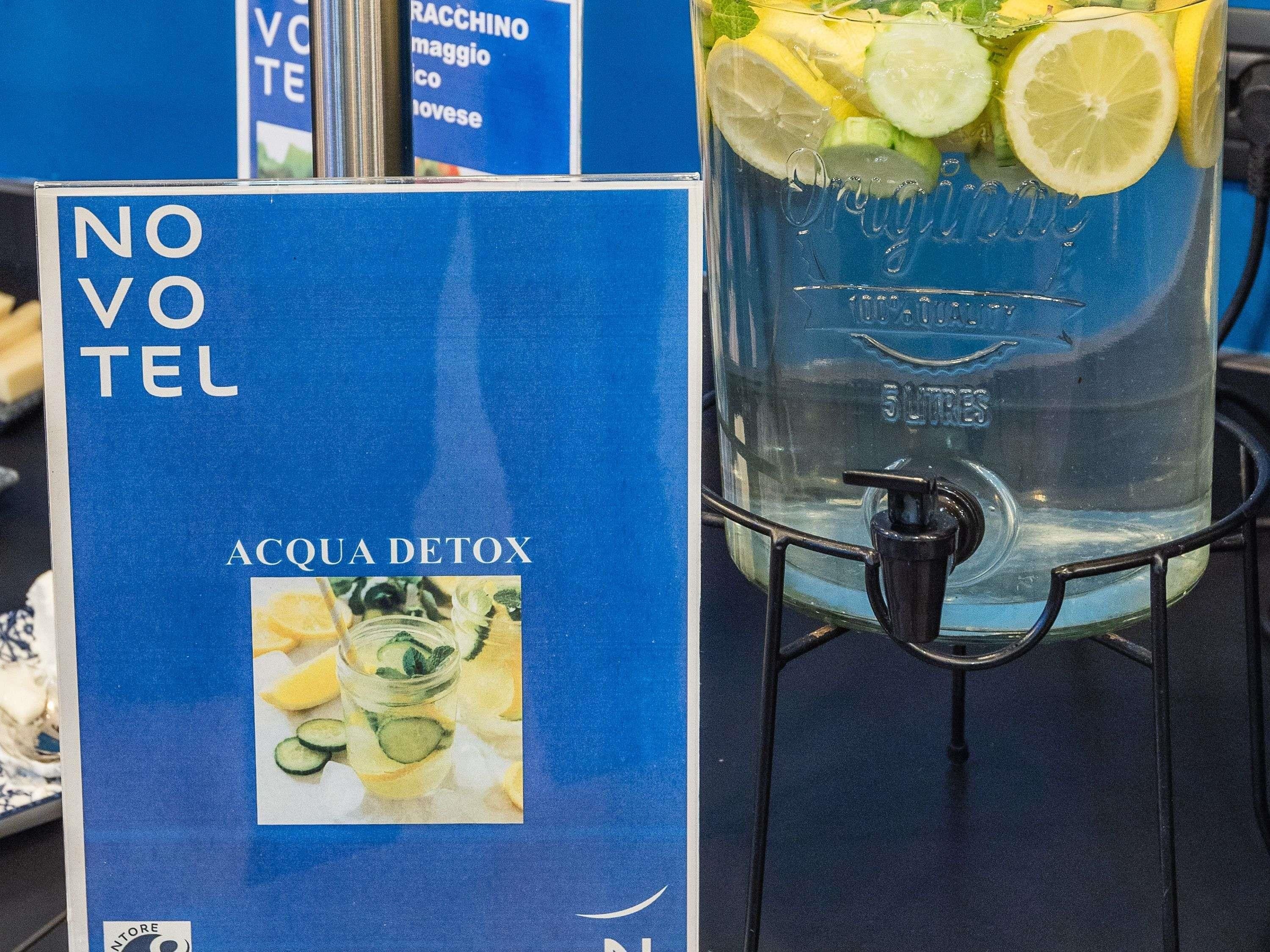 Un distributore d'acqua con un bicchiere pieno d'acqua e fettine di limone, e un cartello con scritto 'NO VOTE ACQUA DETOX'.
