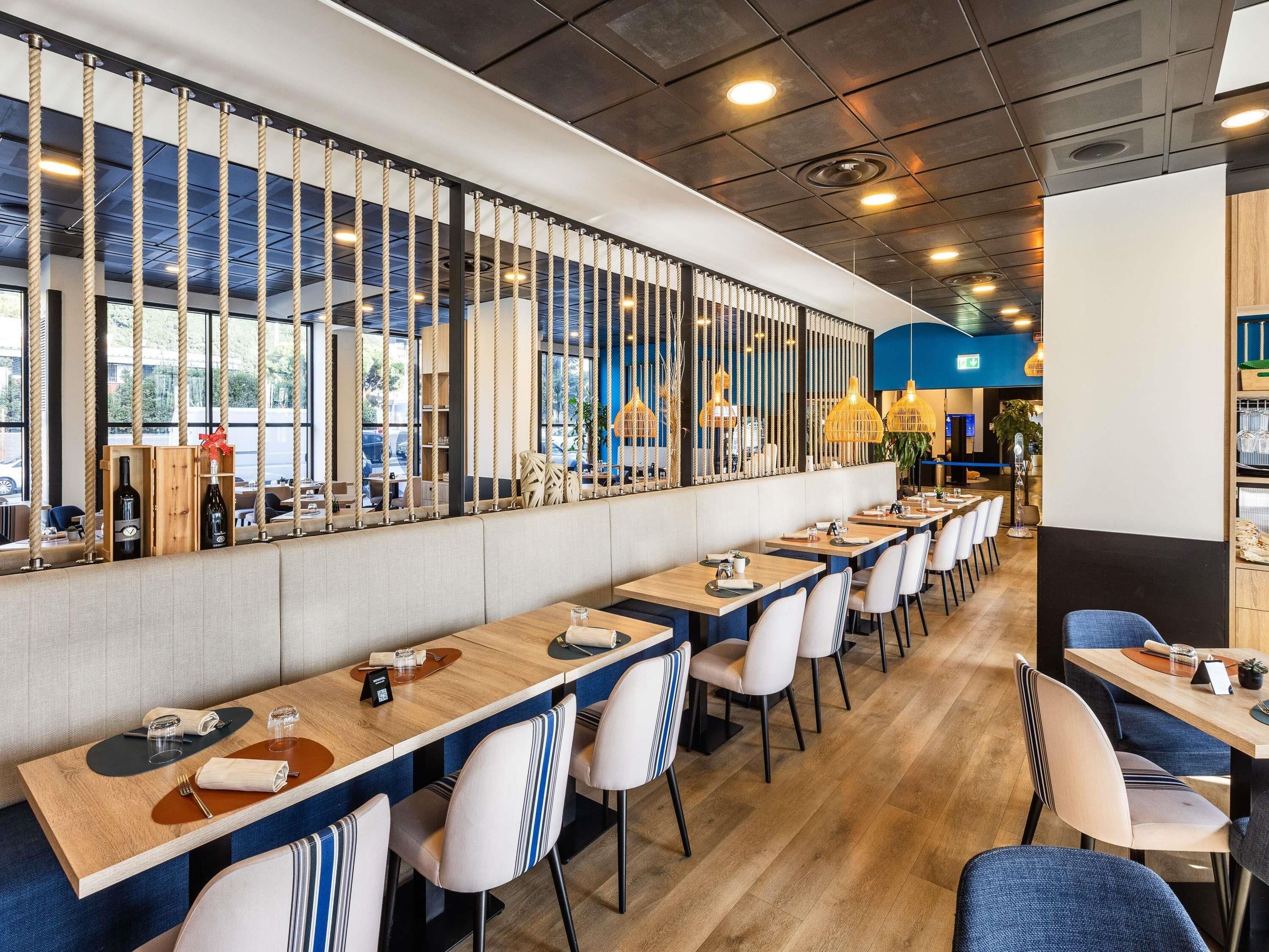 A modern restaurant with long communal tables, high ceilings, and a view of the street.