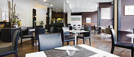 Un restaurante moderno con una distribución limpia y ordenada, con varias mesas y sillas, una zona de cocina visible y un interior bien iluminado.
