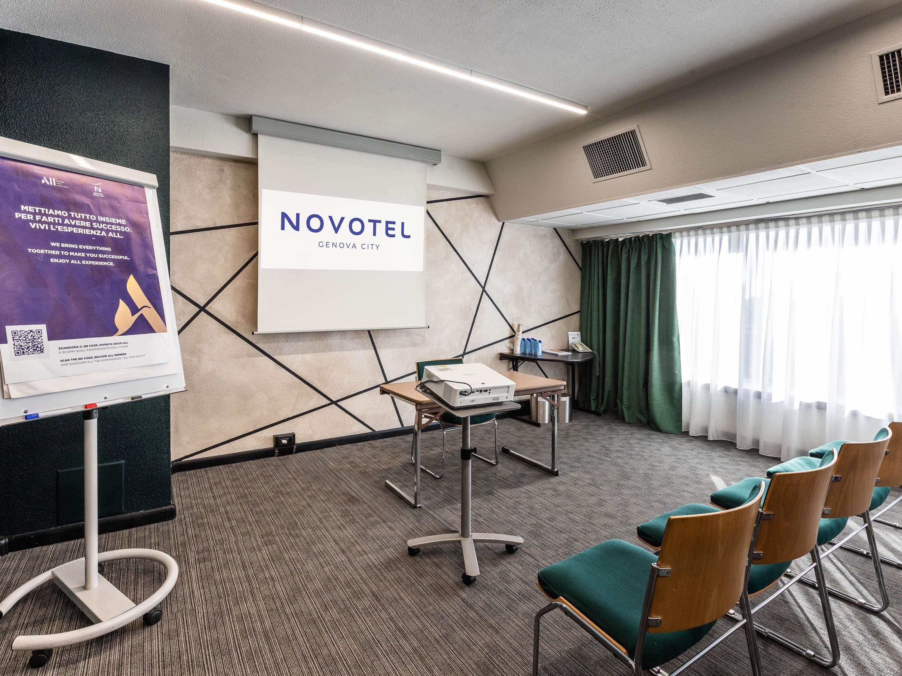 Ein Hotelzimmer mit projizierbarer Leinwand, einem Tisch mit einem Laptop, grünen Stühlen und einem großen Fenster mit Vorhängen.
