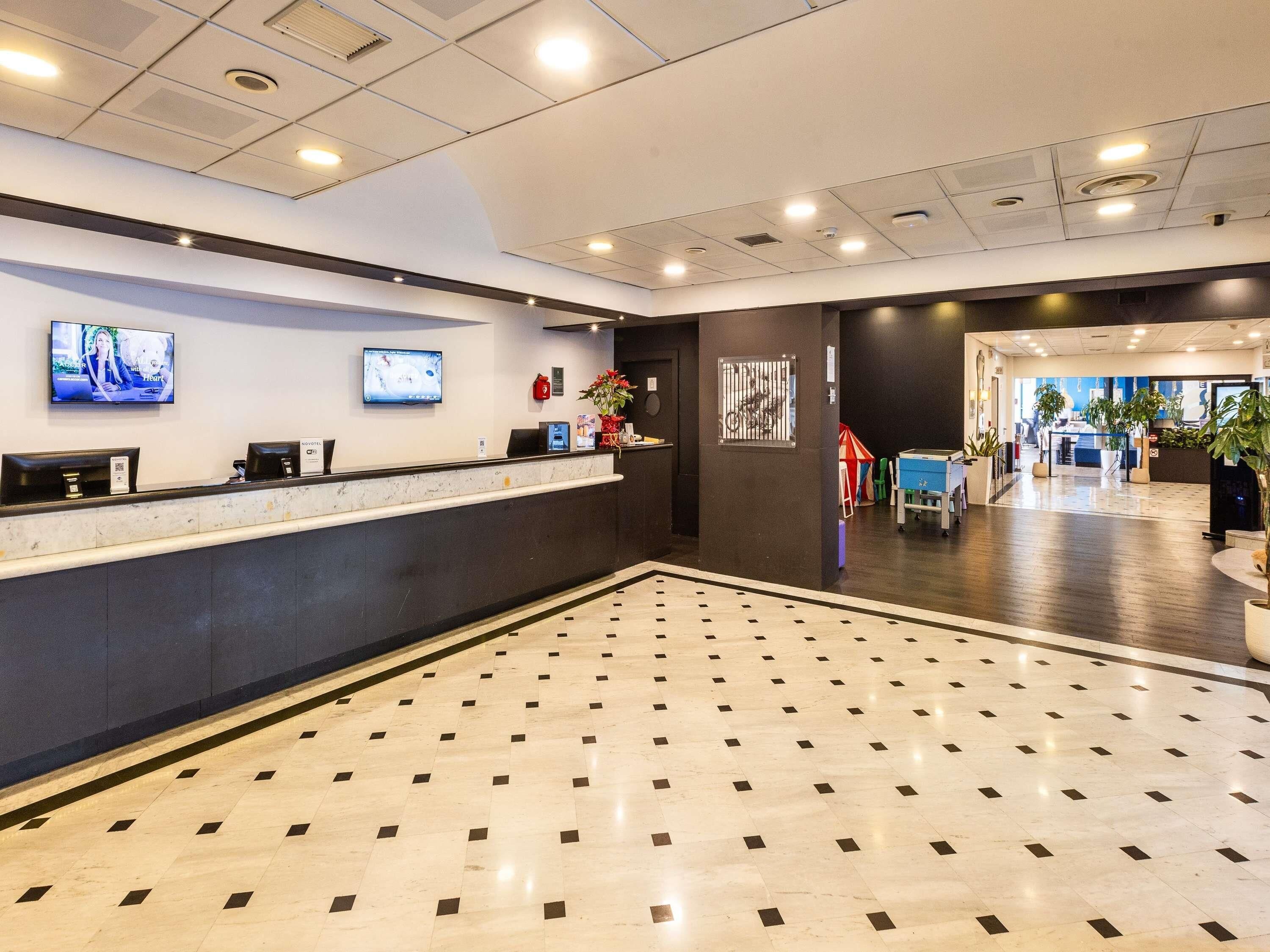 A hotel reception area with a marble floor, two reception desks, a TV screen, and a large window.
