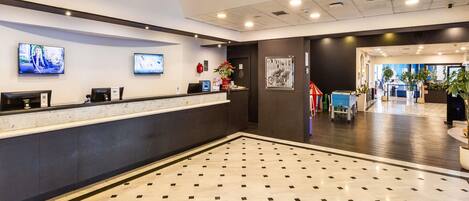 A hotel reception area with a marble floor, two reception desks, a TV screen, and a large window.