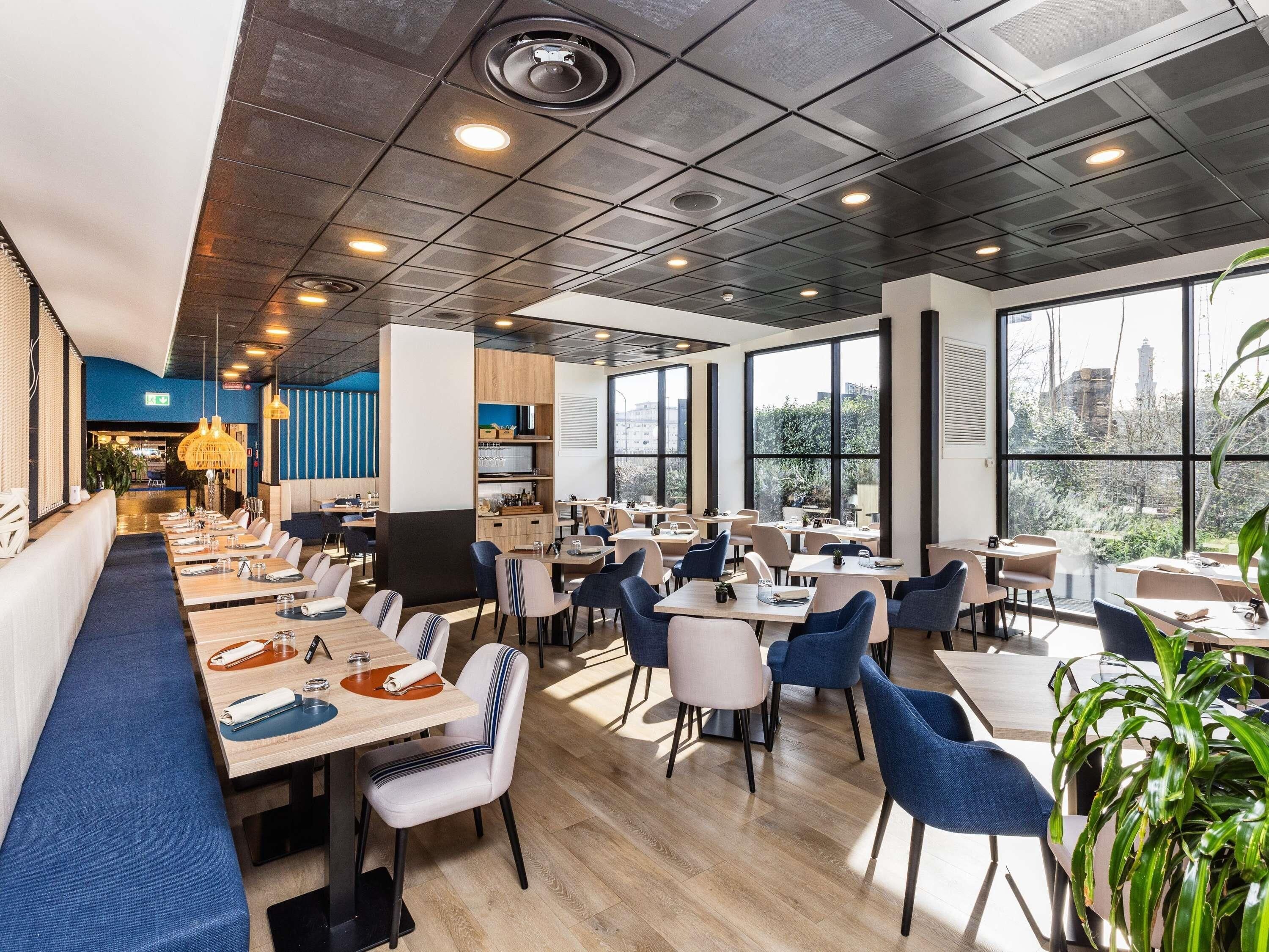 Un restaurant moderne avec un intérieur élégant, caractérisé par de hauts plafonds, de grandes fenêtres et un agencement varié des espaces, comprenant des tables et des banquettes.