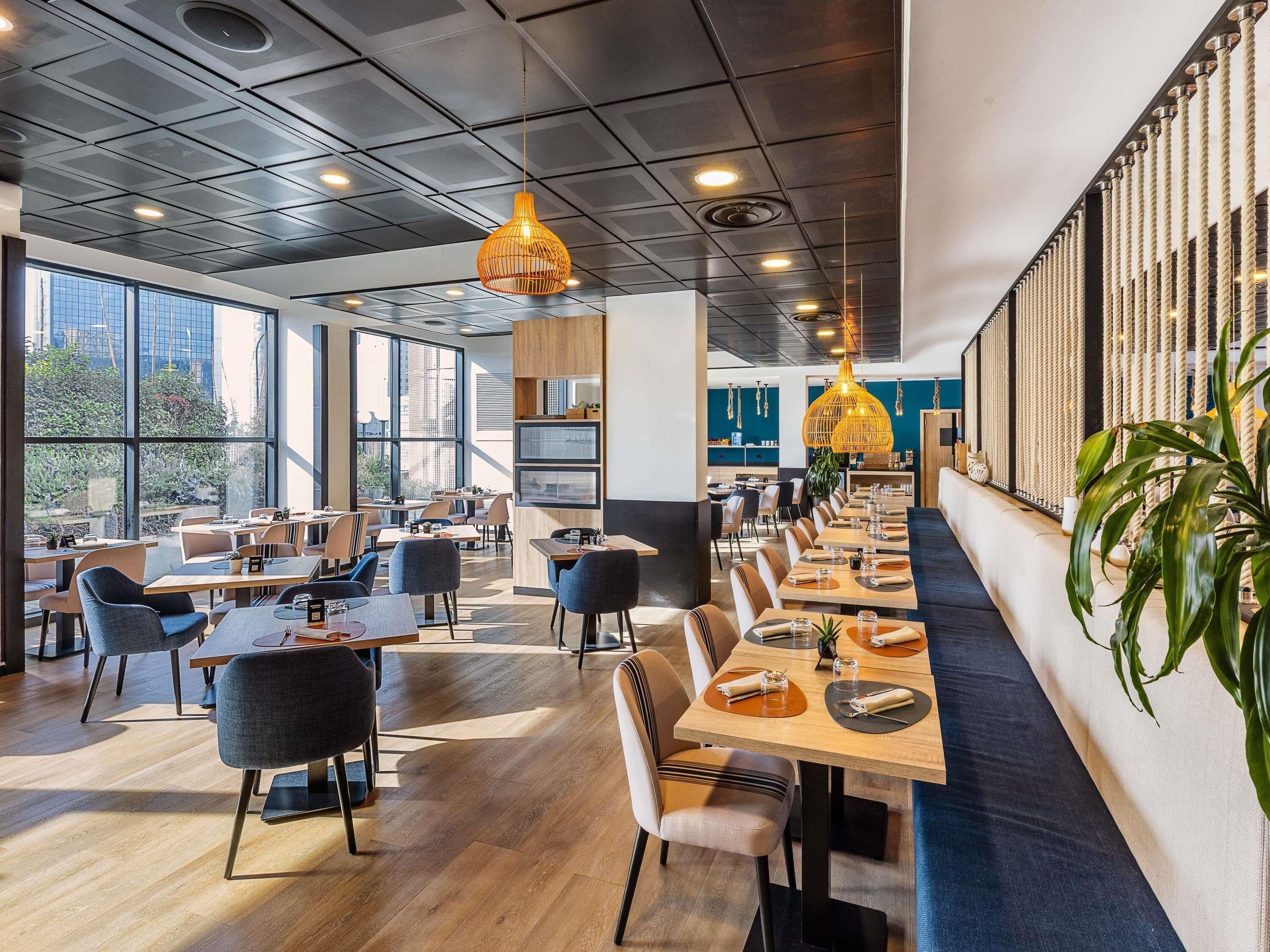 A modern restaurant with long communal tables, high ceilings, and large windows.