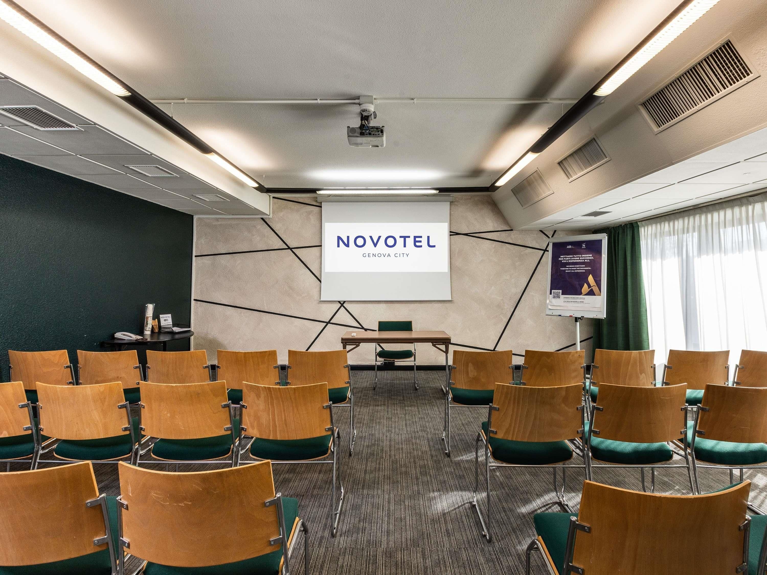 Una sala conferenze con file di sedie e uno schermo proiettore, con una presentazione su Novotel Genova City.