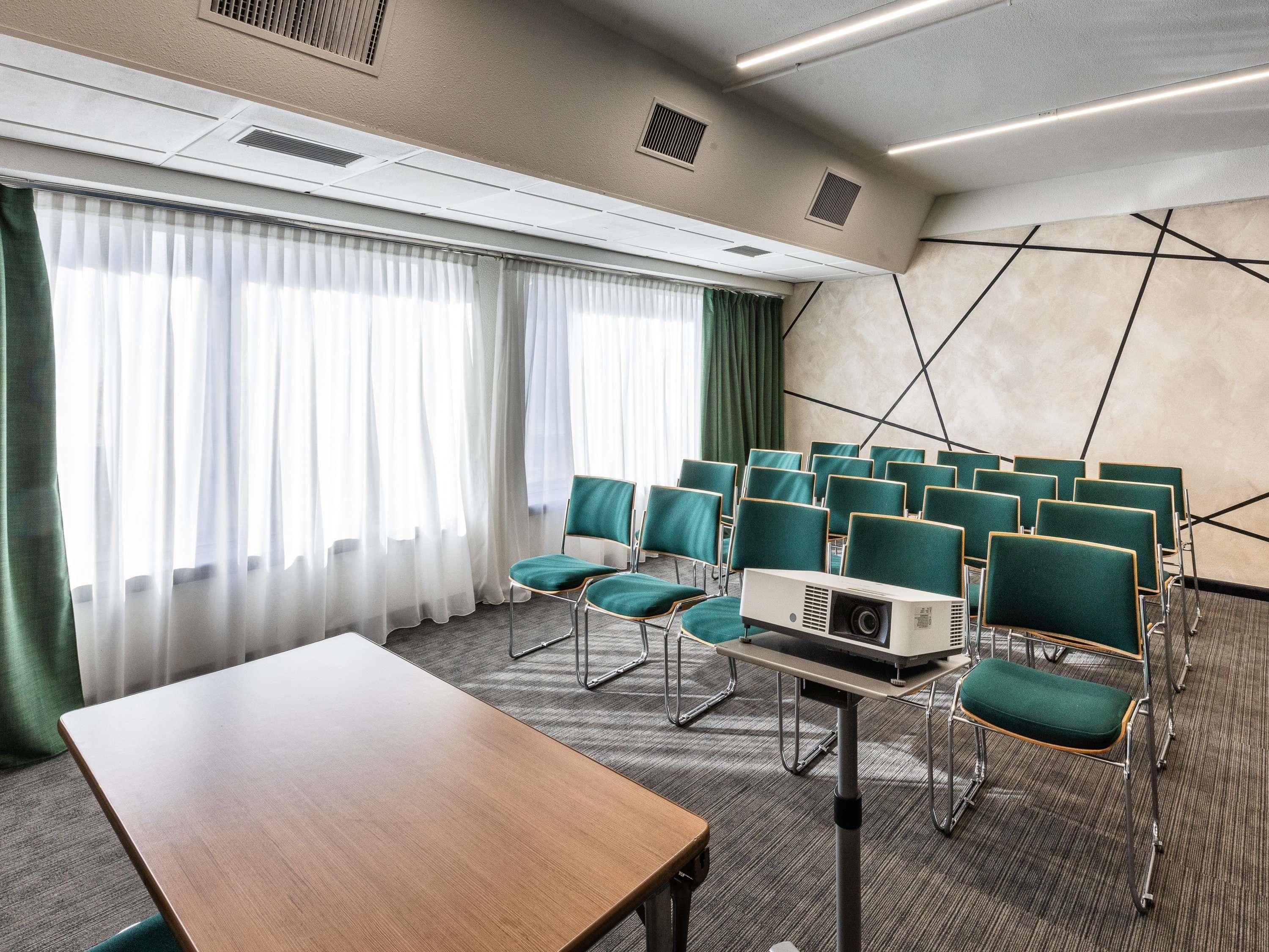 Egy konferenciaterem, amelyben projektor, sorokban elrendezett zöld székek, egy fa asztal és nagy ablakok találhatók, melyekre áttetsző függönyök vannak függesztve.