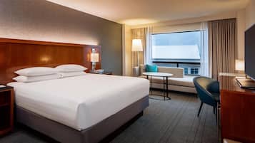 Quarto de hotel com uma cama grande, uma escrivaninha, uma cadeira, uma mesinha e uma janela com cortinas.