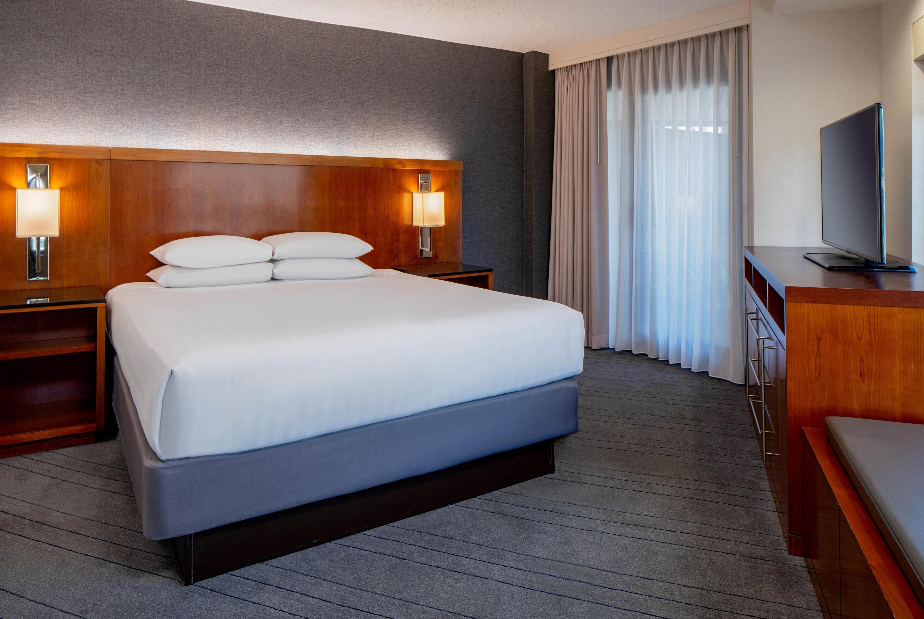 Une chambre d’hôtel avec un grand lit, deux tables de chevet avec des lampes, un téléviseur posé sur une commode en bois, et une vue sur l’extérieur par la fenêtre.
