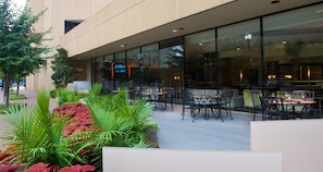 Un área de comedor al aire libre con mesas y sillas, rodeada de follaje y la fachada de un edificio.