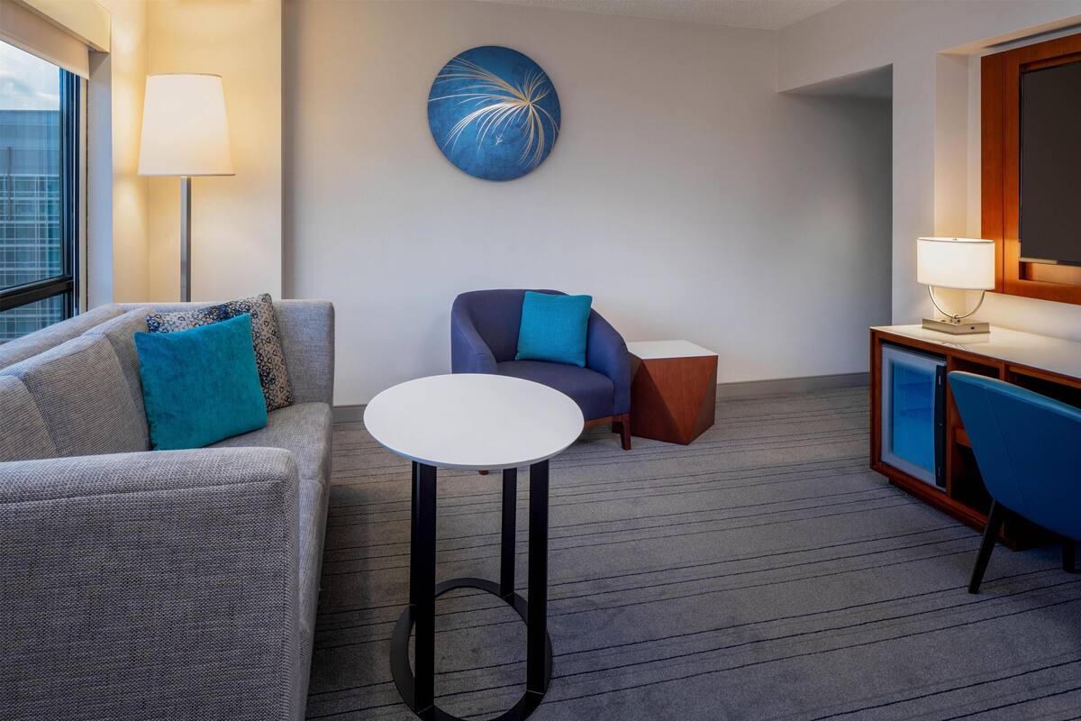 Una habitación de hotel con sofá, una mesita redonda, un sillón azul, un armario y una obra de arte en la pared.