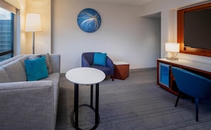 Una habitación de hotel con sofá, una mesita redonda, un sillón azul, un armario y una obra de arte en la pared.