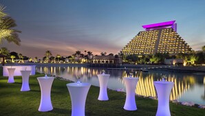 Un hotel moderno con una estructura piramidal distintiva, un lago tranquilo y mesas blancas iluminadas con flores.