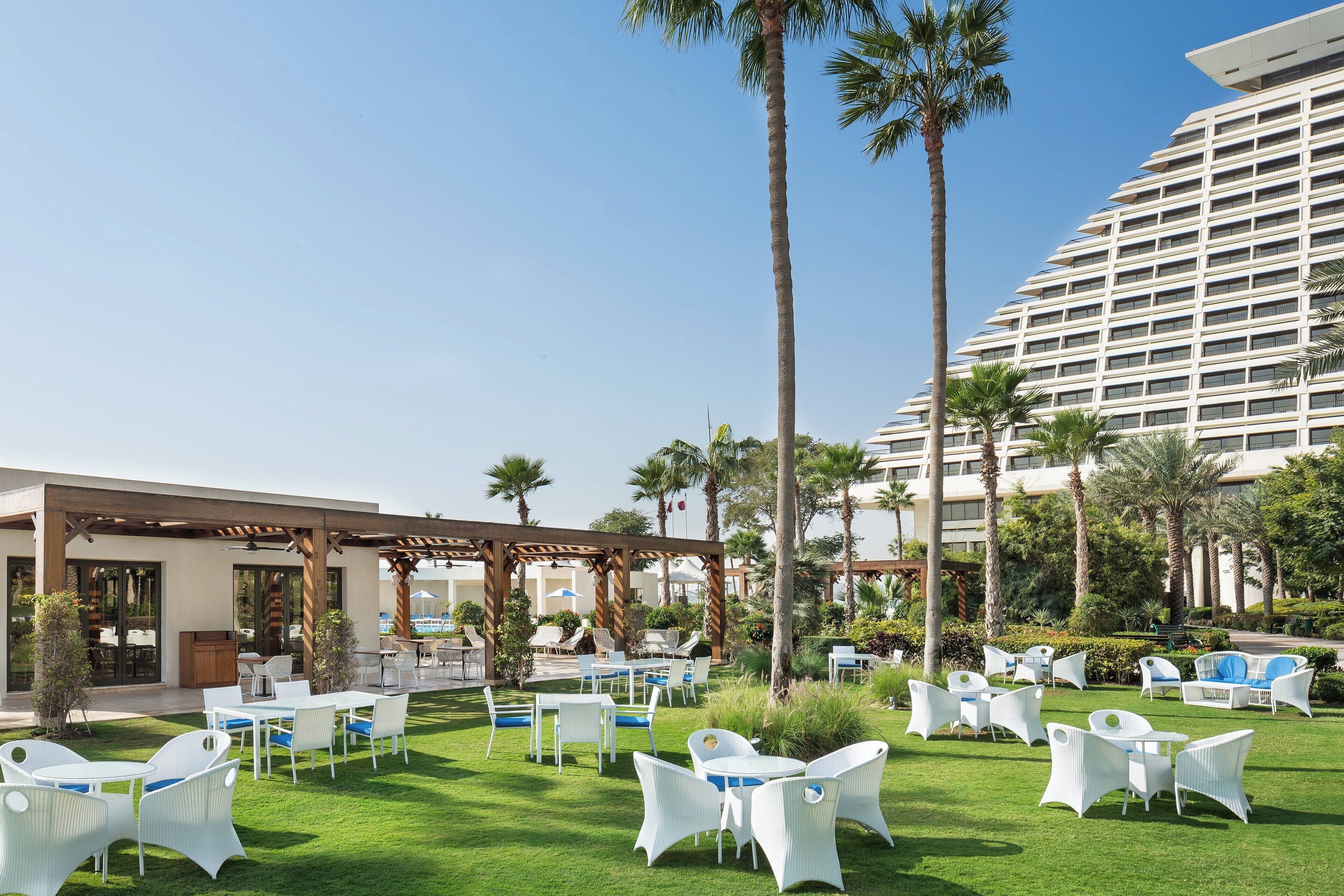 An outdoor patio with white wicker furniture, palm trees, and a modern building in the background.