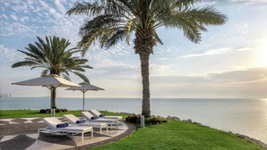 Zona junto a la piscina con sillones de descanso y sombrillas, una palmera y vista al mar.