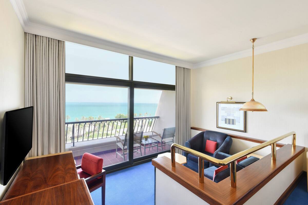 A hotel room with a large window, a flat-screen TV, a wooden desk, a sofa with cushions, and a hanging lamp.