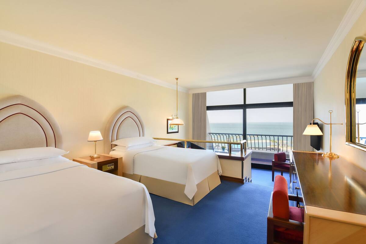 A hotel room with two beds, a desk, a chair, and a view of the sea.