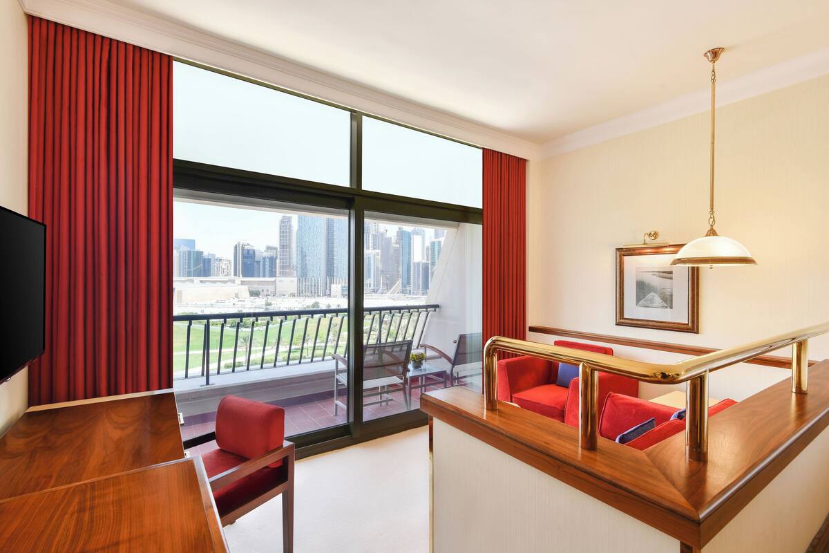 A hotel room with a balcony, a wooden desk, red seating, and a city view.
