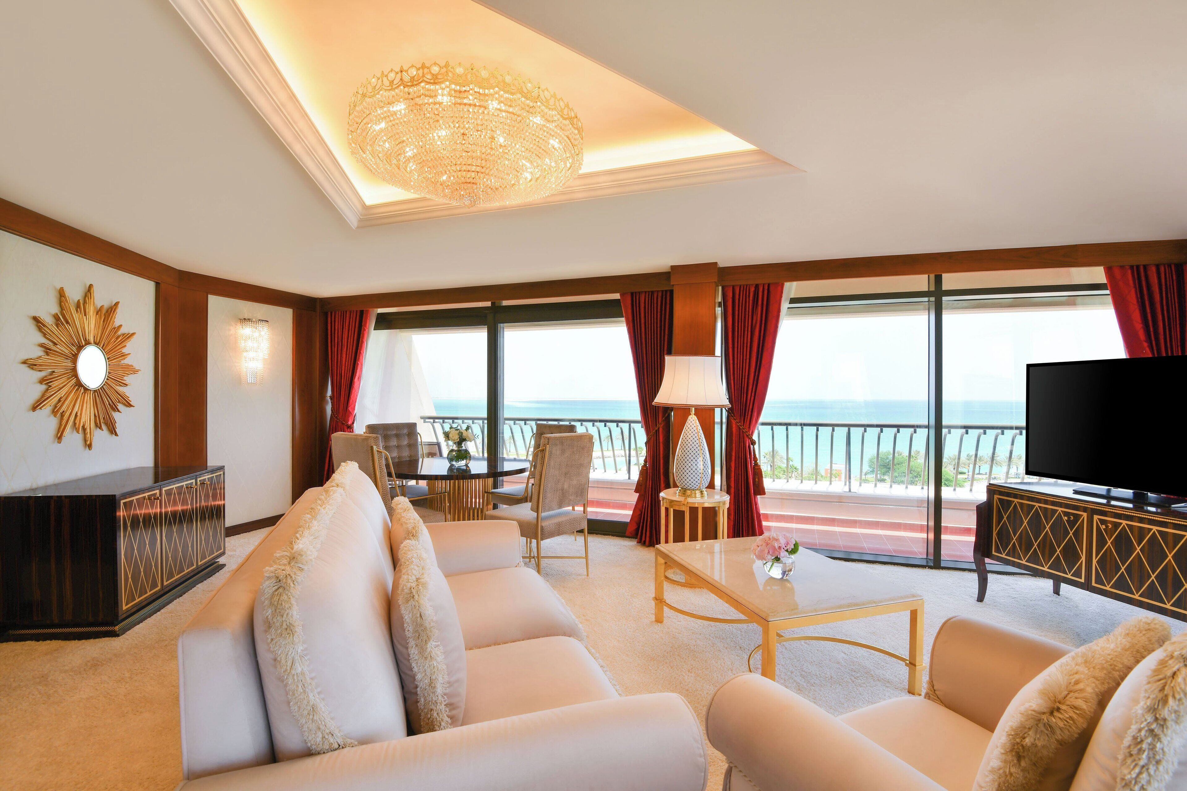 A spacious living room with a large sofa, a coffee table, and a television. There is a balcony with a view of the ocean.